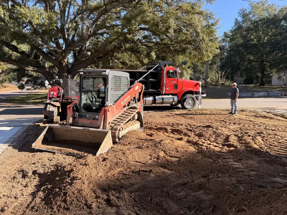 Red dump truck and track loader on a dirt lot under a live oak